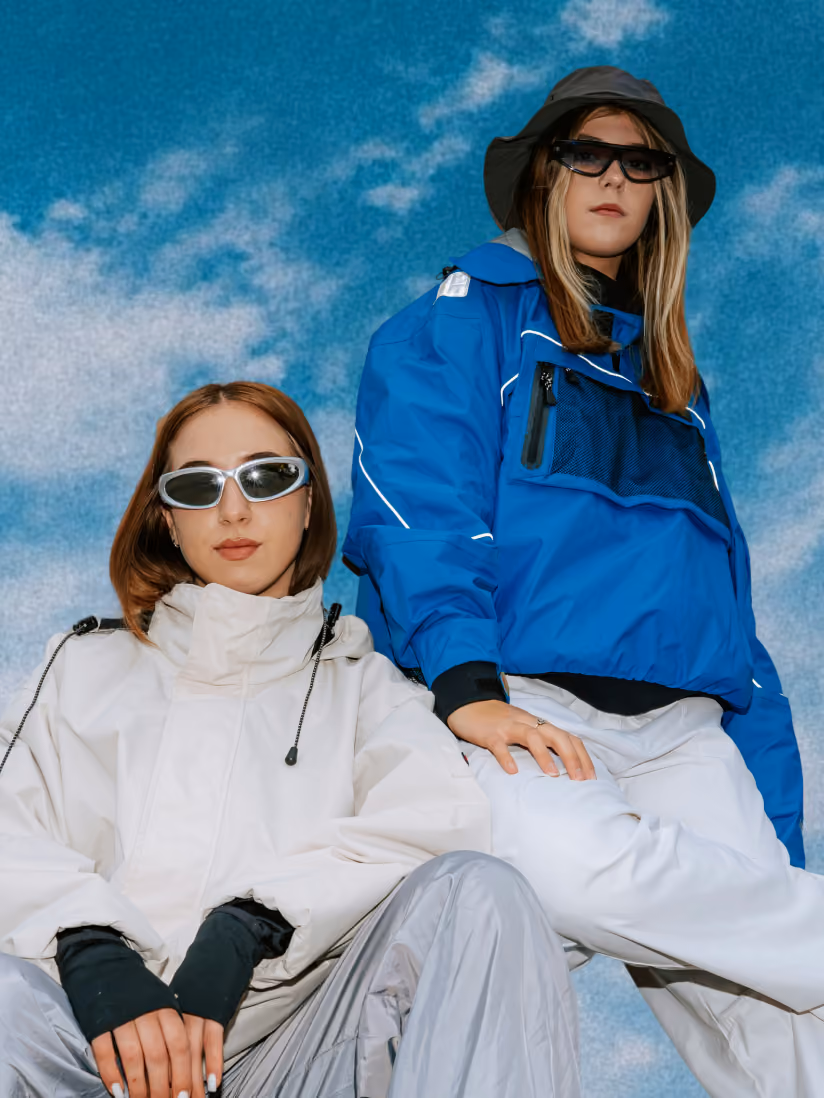 Two women wearing techwear jackets and sunglasses pose against a blue sky with clouds.