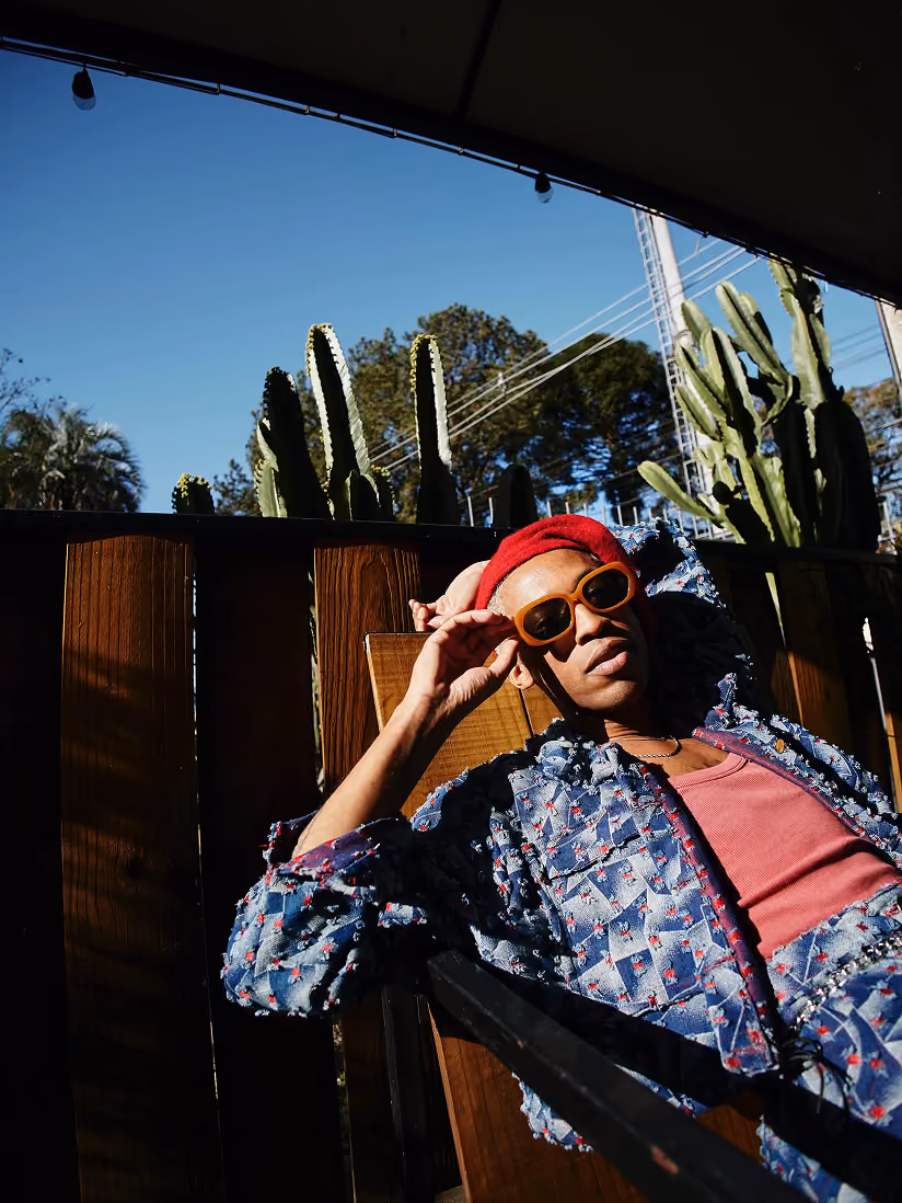 Person in a red beanie and sunglasses reclining outdoors beside tall cacti under a clear blue sky.