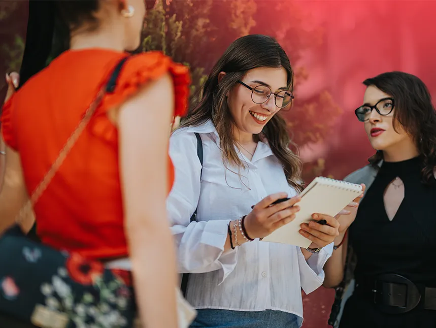 A smiling young woman with glasses and long brown hair wearing a white button-up shirt looks down at a small notebook in her hands, surrounded by two other women in a warm, social setting with reddish background tones.