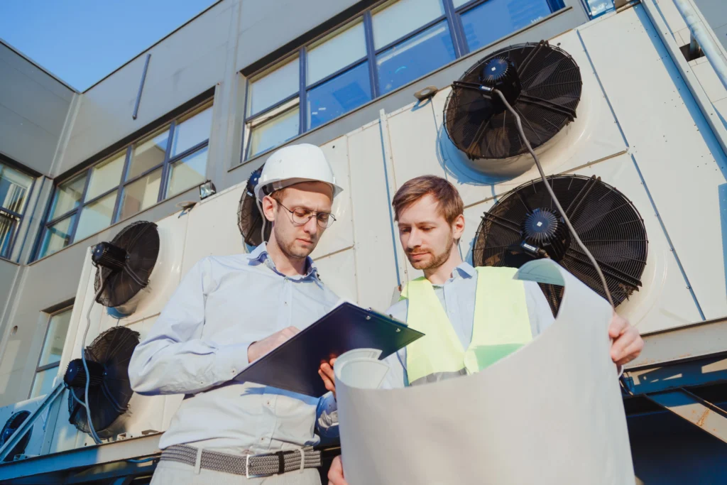HVAC engineers reviewing plans in front of industrial air conditioning units, highlighting modern HVAC systems and maintenance practices.