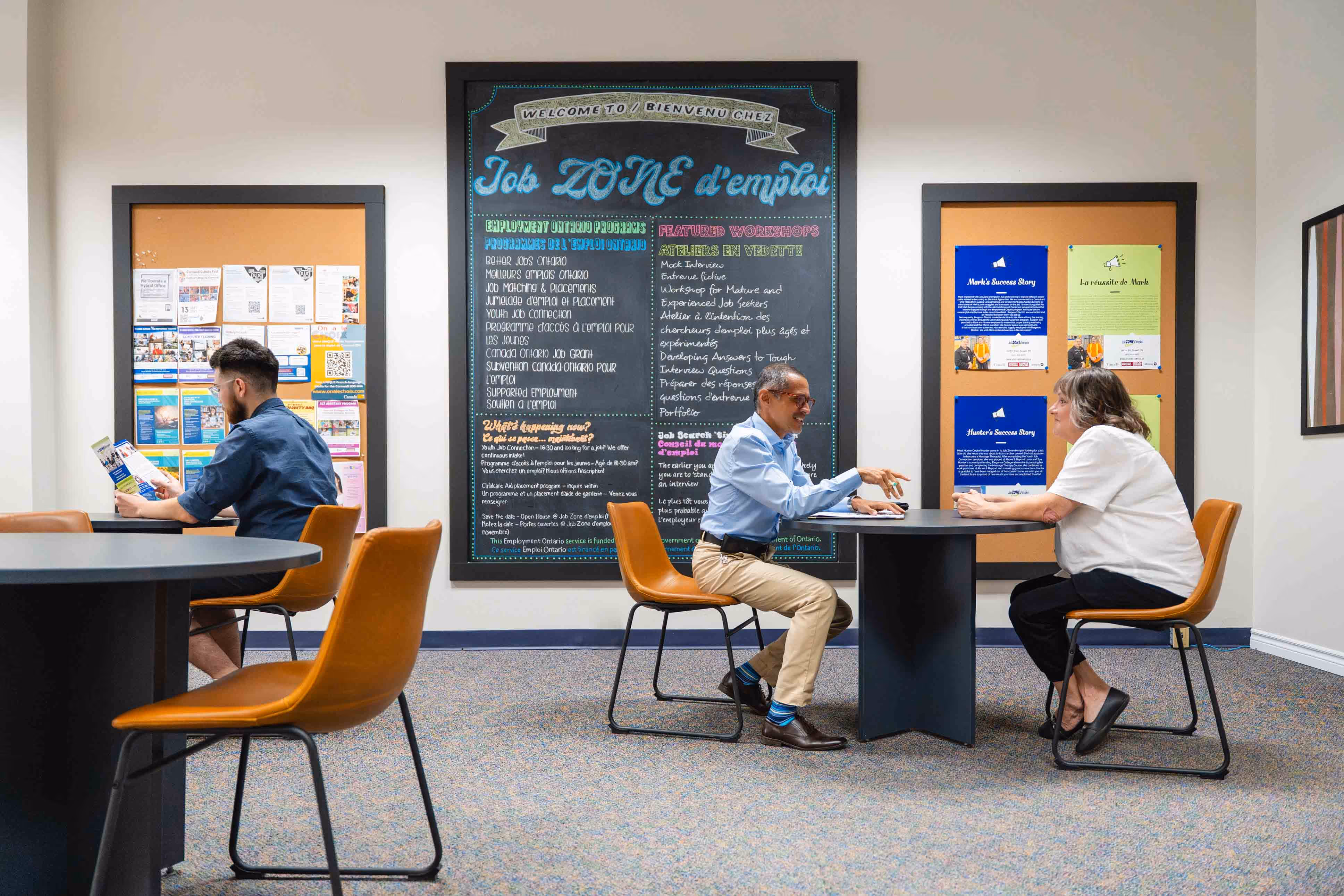 Two people seated at a table in a job resource centre with another man reading flyers nearby.
