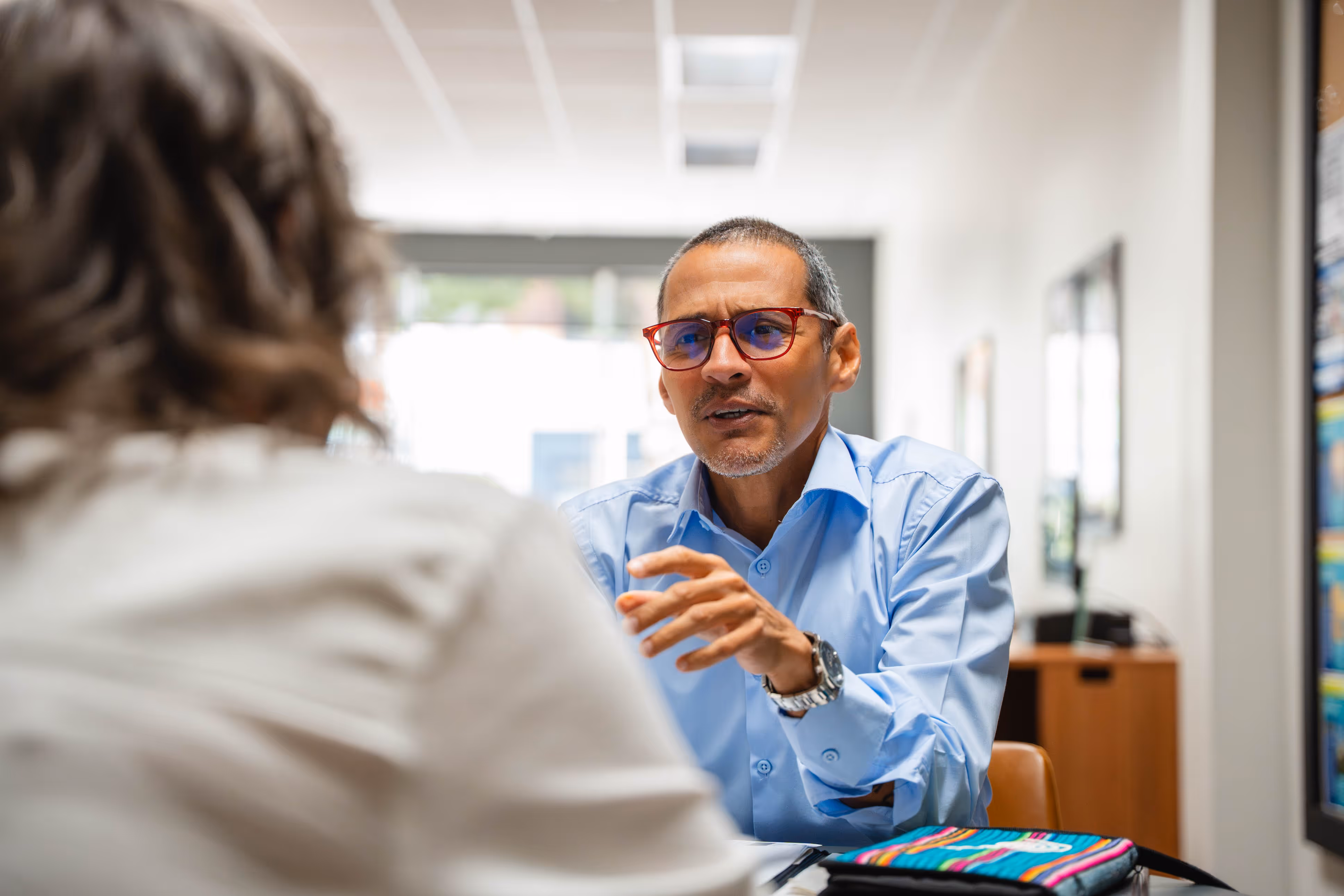 Man in red glasses and blue shirt having a focused conversation with a woman at a table in an office.