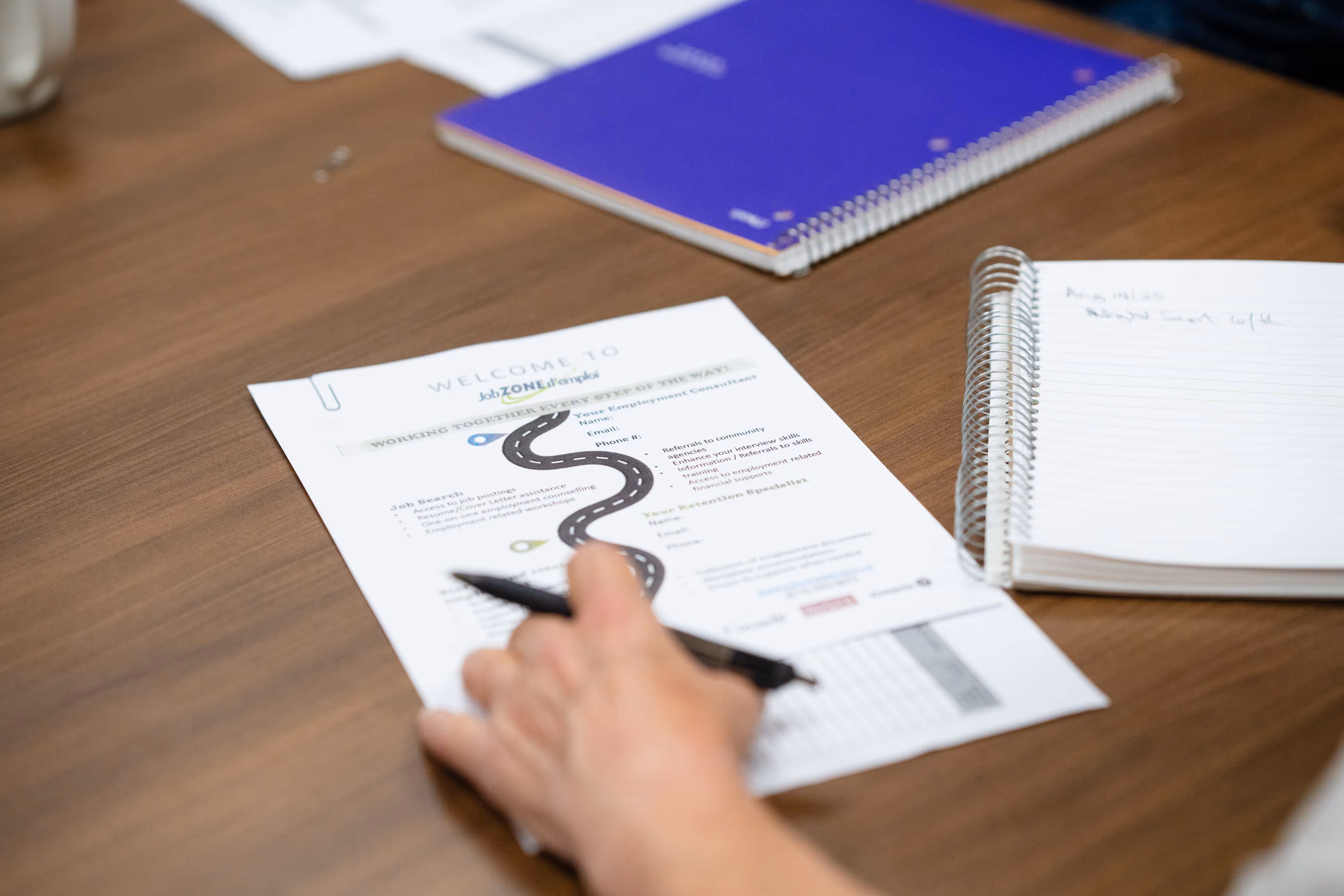 Hand holding a pen over a Job Zone d'emploi employment assistance flyer on a wooden desk, next to a spiral notebook with handwritten notes and a purple notebook.