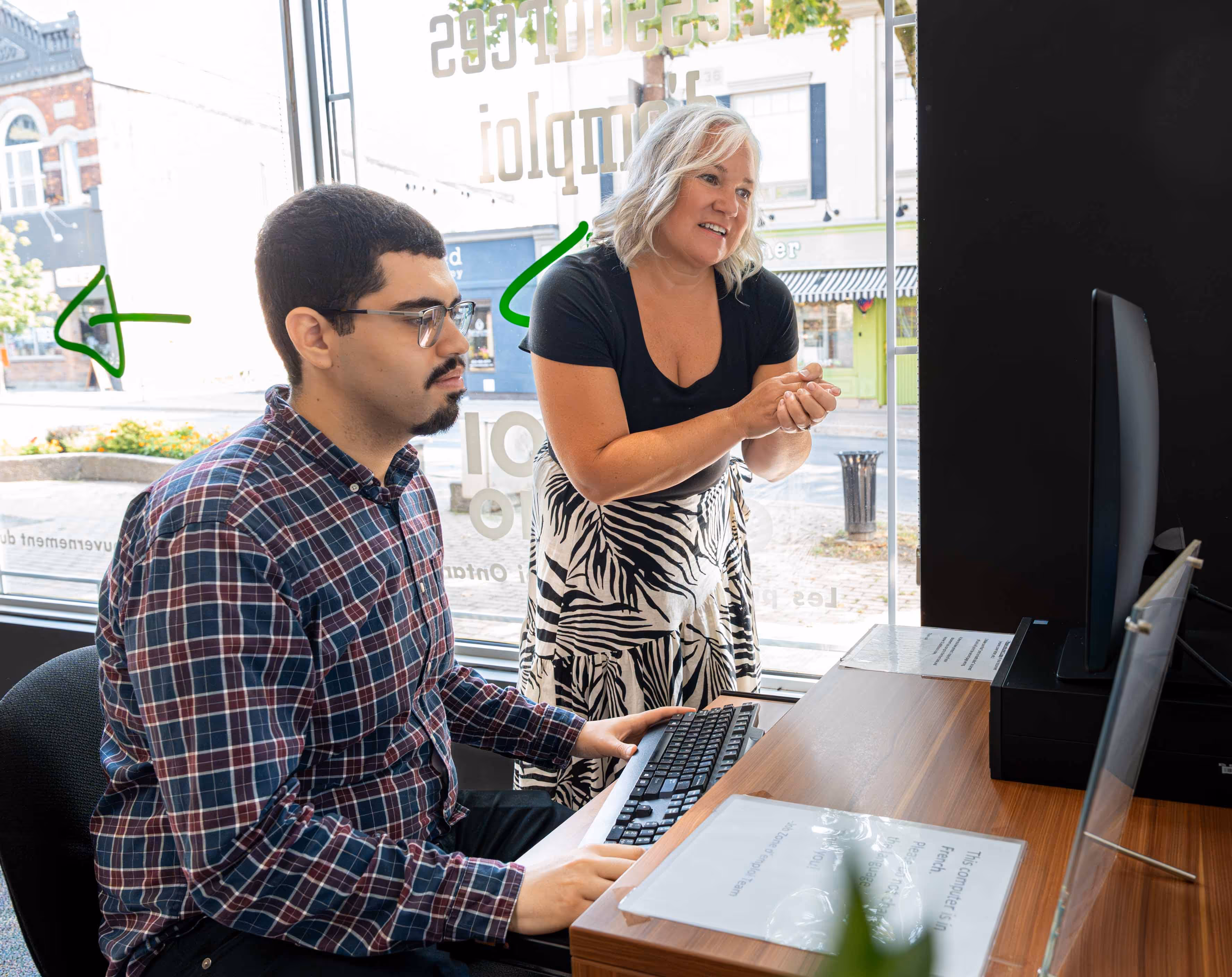Woman standing and smiling while assisting a seated man who is typing on a keyboard at a desk with a computer monitor indoors.