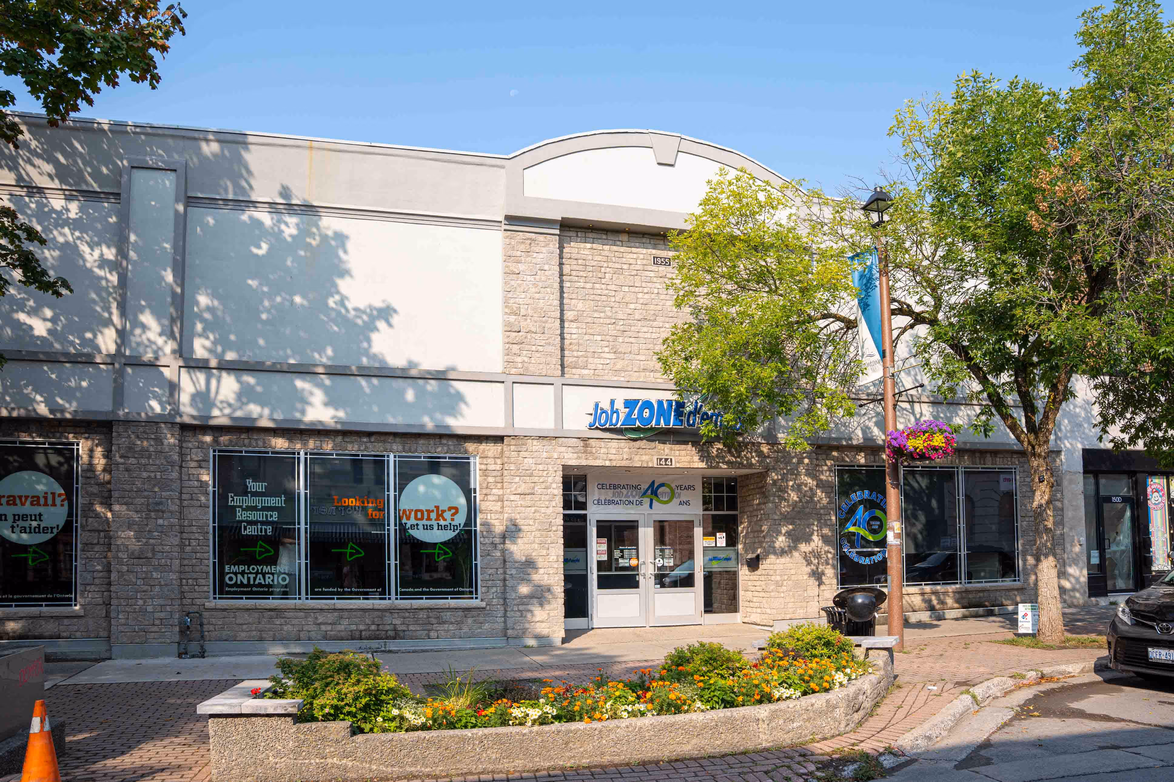 Exterior of Job Zone d'emploi employment resource centre with stone façade, glass entrance doors, and flower bed in front.
