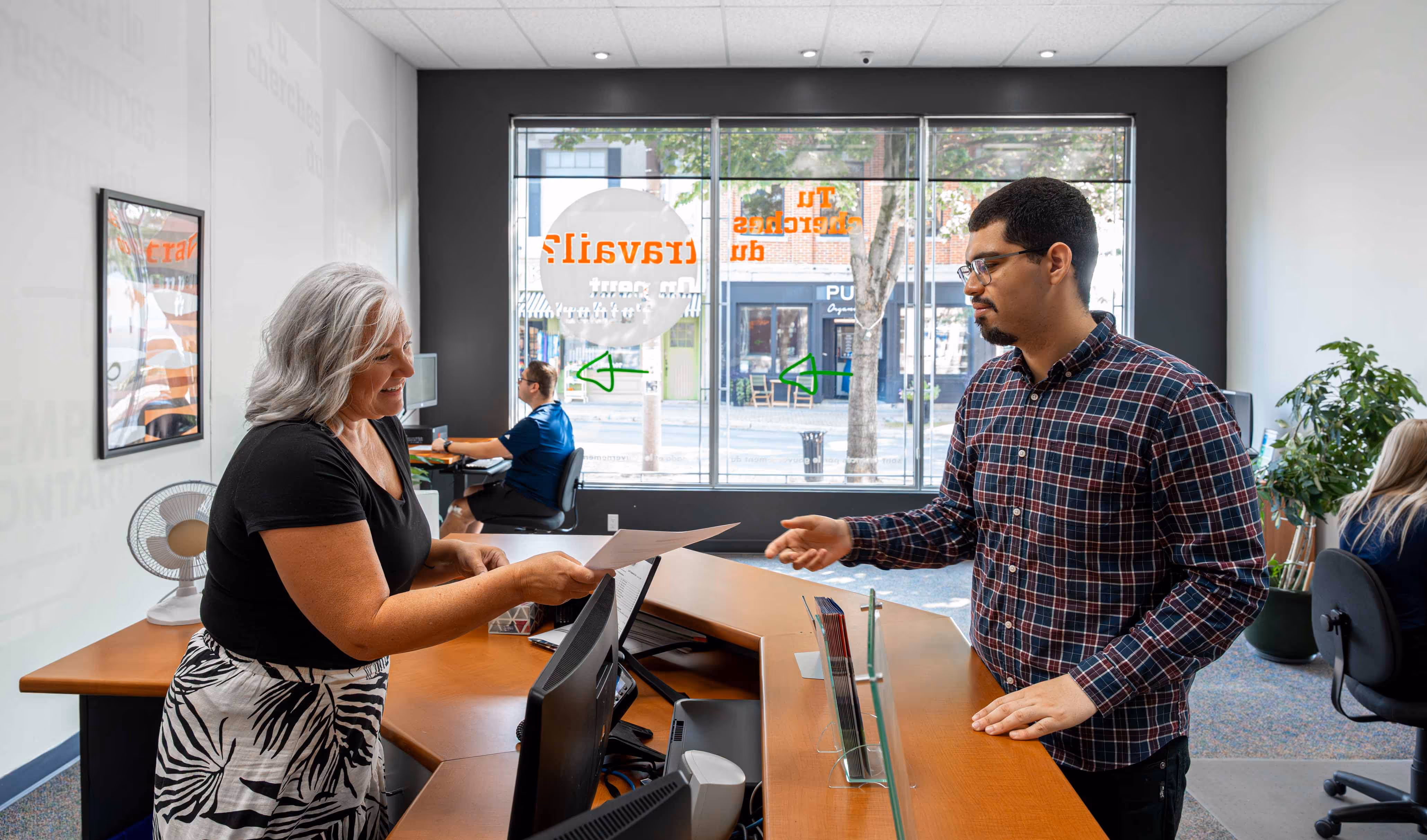 Smiling woman hands paper to man across office reception desk in a bright modern workspace.