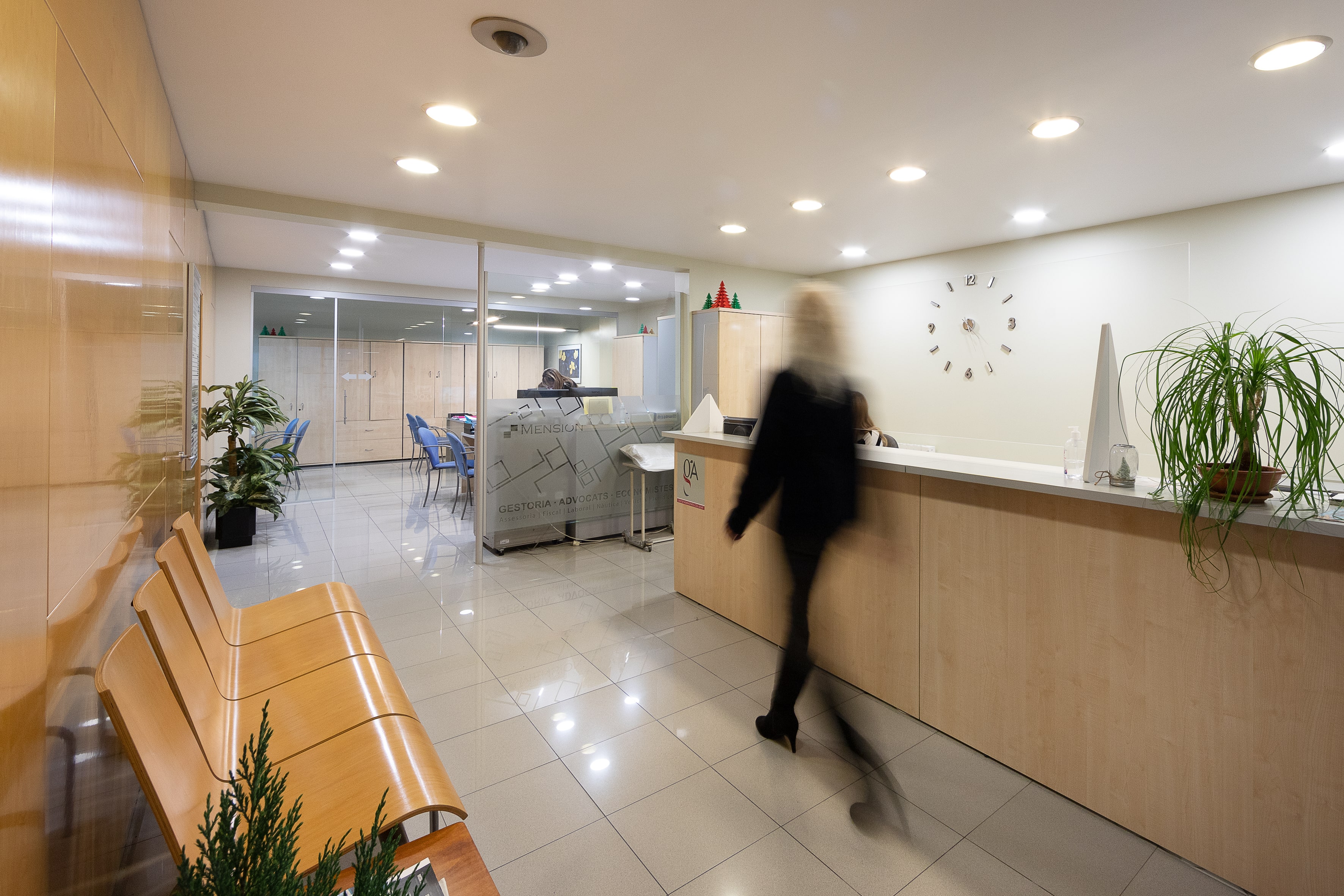 Recepción de oficina moderna con mostrador de madera, planta en maceta, sillas de espera y una persona borrosa caminando.