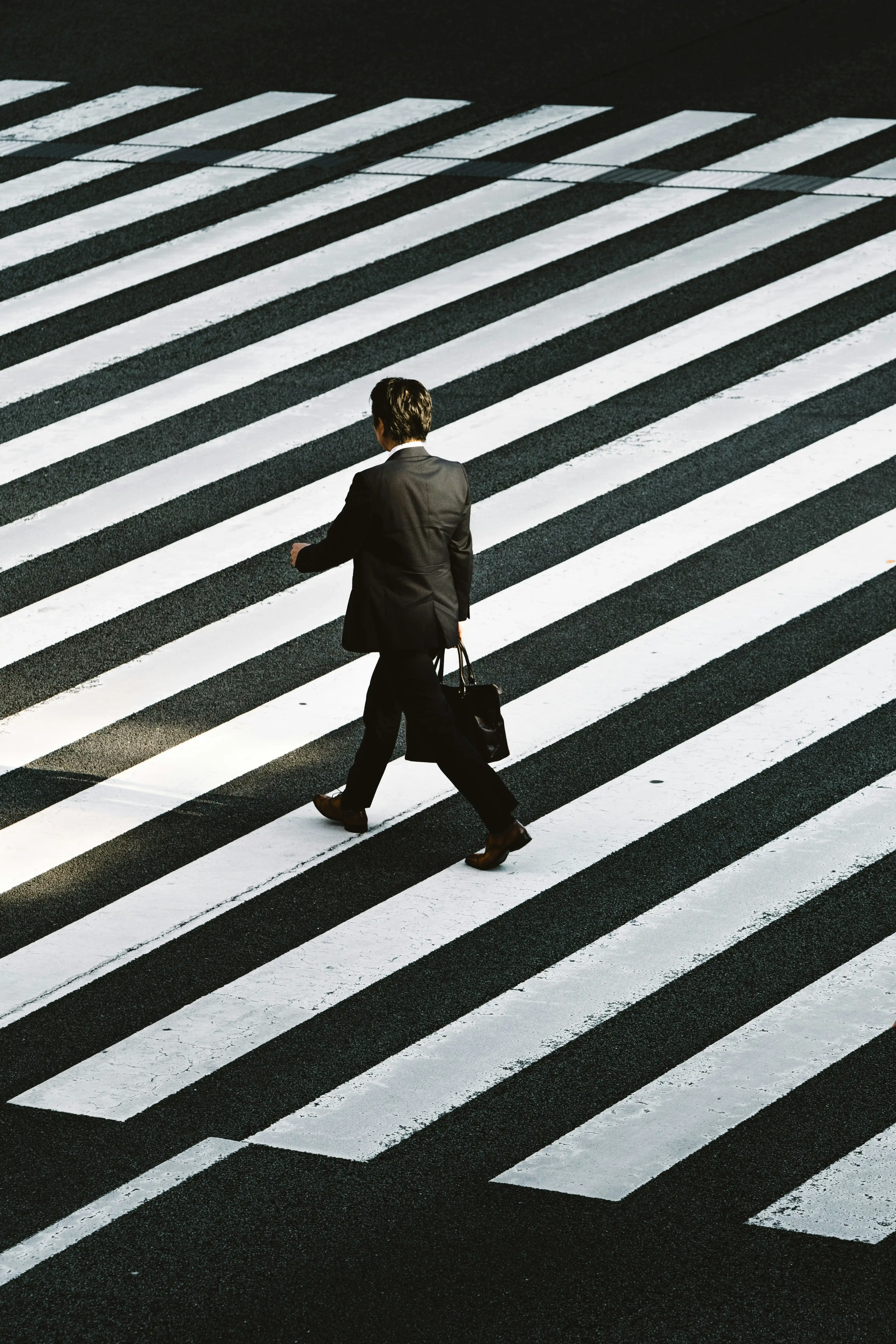 Hombre de negocios caminando con traje negro y portafolios sobre un paso peatonal con franjas blancas y negras.