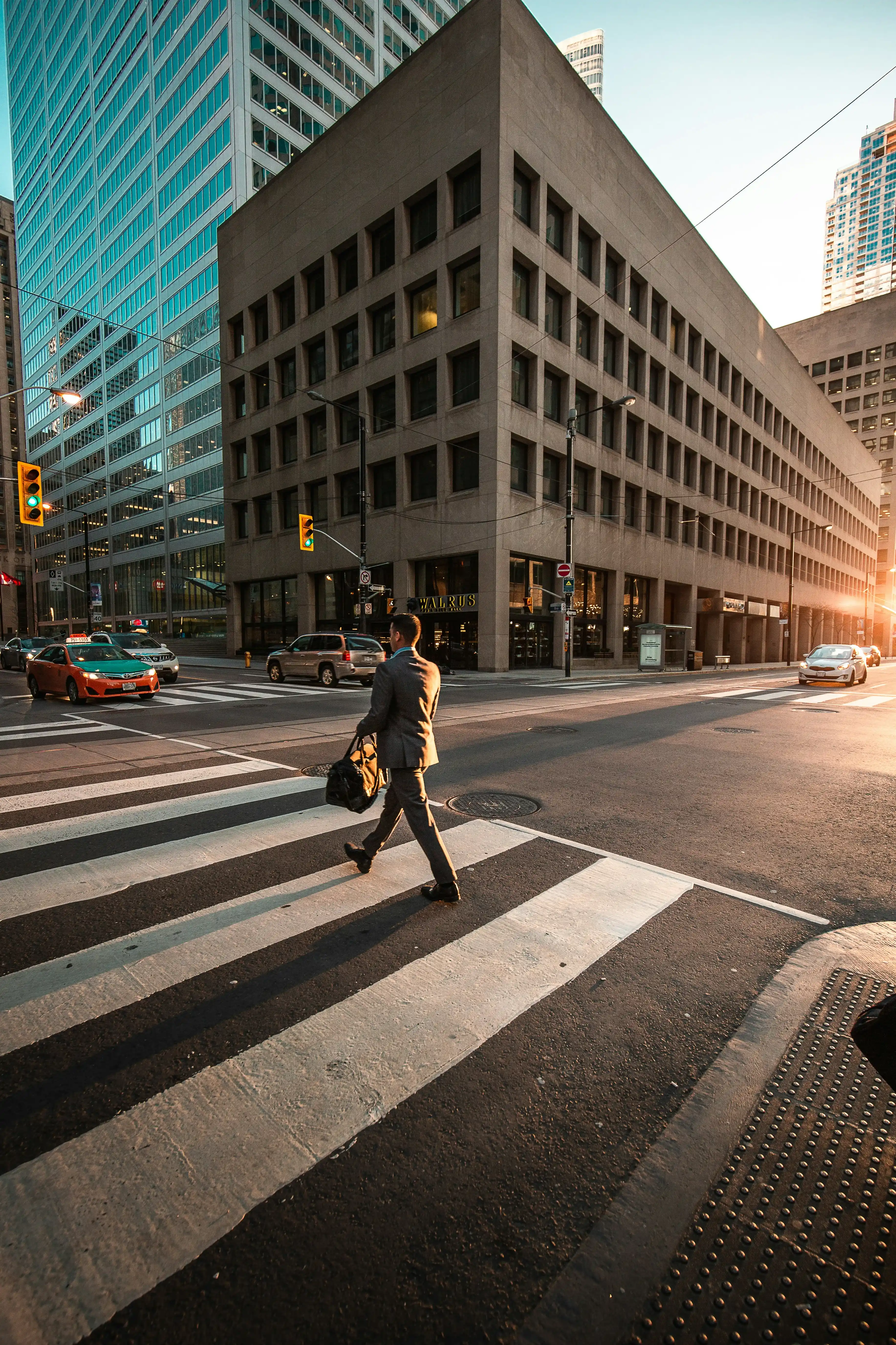 Hombre con traje cruzando un paso de peatones en una intersección urbana al atardecer con autos y edificios altos.