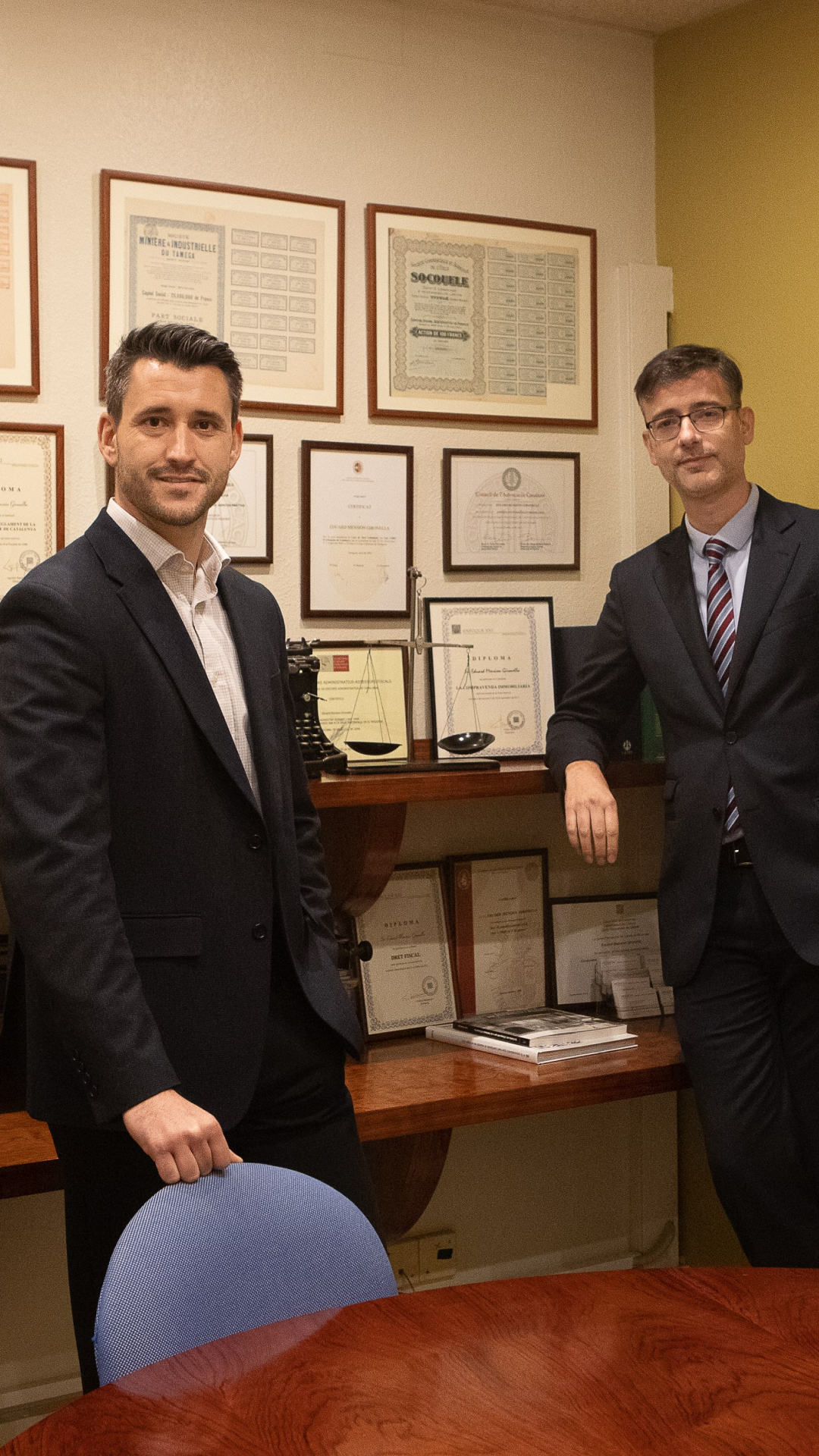 Dos hombres de negocios vestidos formalmente posan en una oficina con diplomas y certificados en la pared.