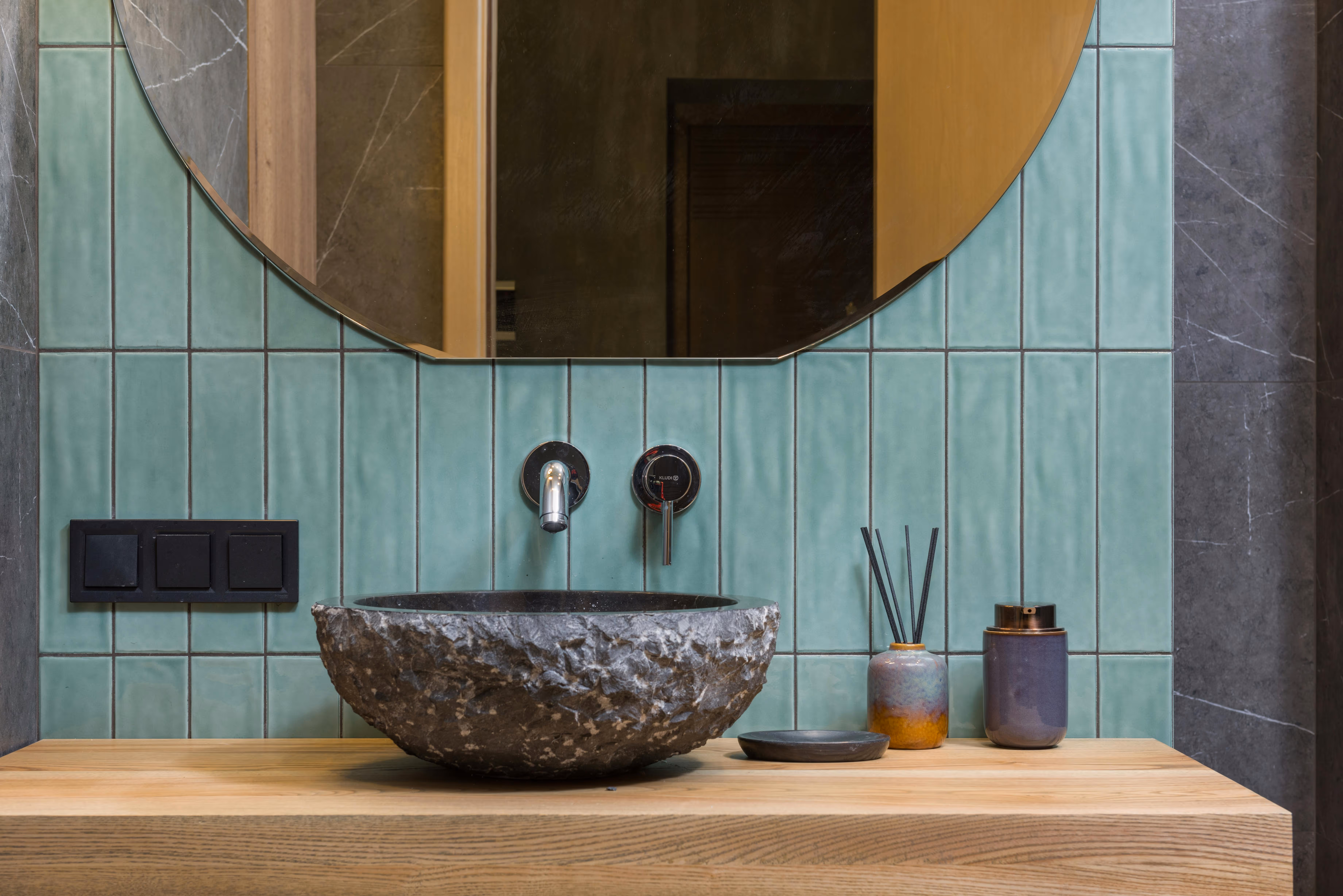Modernes Badezimmer mit grünen Fliesen, rundem Wandspiegel und einem Steinwaschbecken auf einer Holzablage