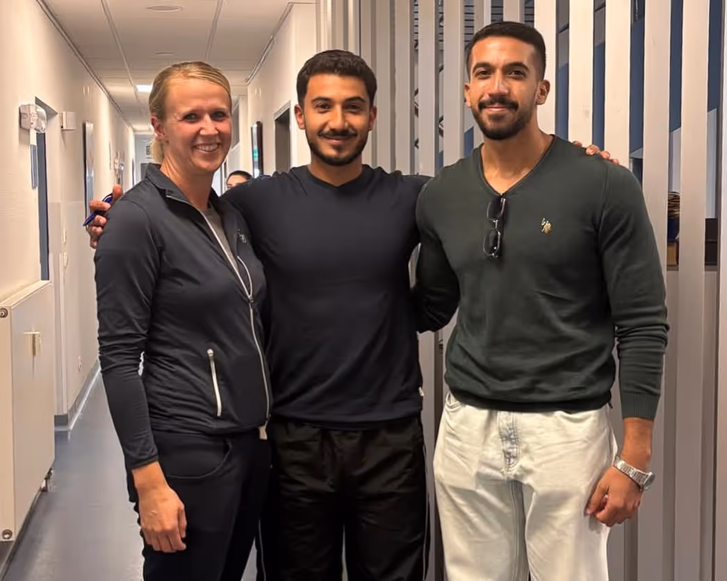 Drei lächelnde Personen, zwei Männer und eine Frau, stehen in einem Flur zusammen und umarmen sich.