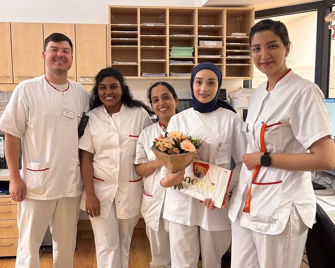 Fünf medizinische Fachkräfte in weißer Arbeitskleidung lächeln zusammen, eine hält Blumenstrauß und Dankeschön-Schokolade.