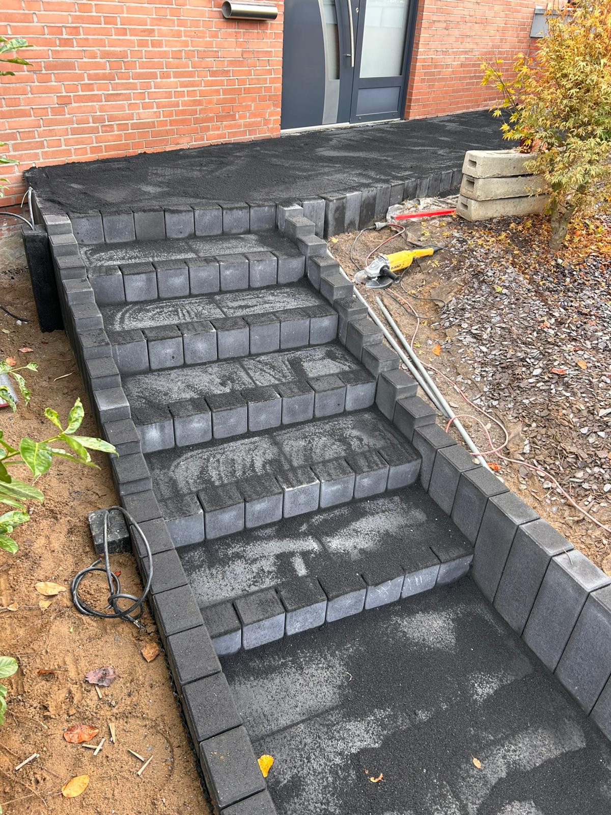 Unfertige graue Blockstufen mit schwarzem Sand vor einem roten Backsteinhaus mit grauer Tür.