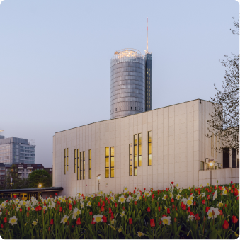 Moderne Stadtansicht mit einem hohen runden Gebäude im Hintergrund und einem weißen Gebäude mit gelben Fenstern sowie Blumen im Vordergrund.