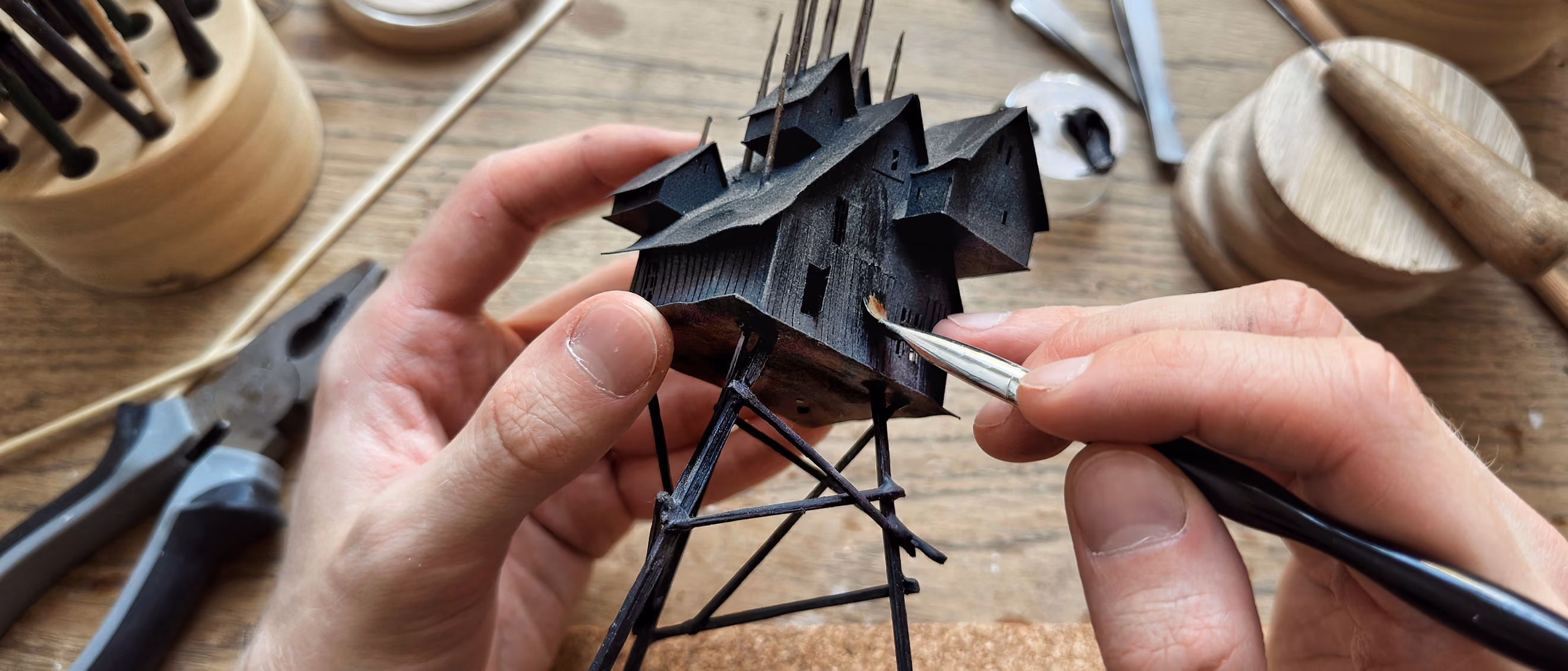 Close-up of hands carefully painting a small, detailed black model structure resembling a house on stilts with a fine brush, on a wooden workbench with pliers and other tools nearby.