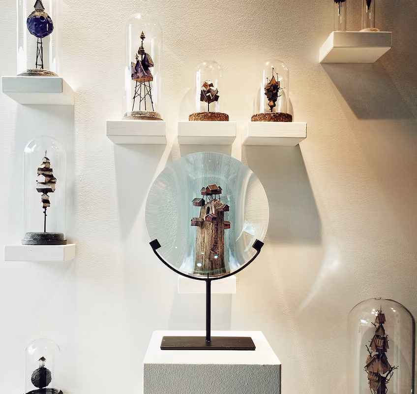 Display of small wooden sculptures of abstract towers or houses encased in glass domes on white wall-mounted shelves and a pedestal, with one central sculpture magnified behind a large round glass lens on a stand.