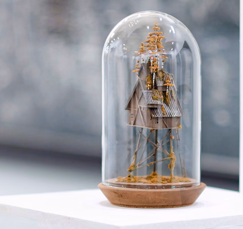Miniature wooden house with a slanted roof and small trees on top, elevated on thin stilts, enclosed inside a clear glass dome with a wooden base.