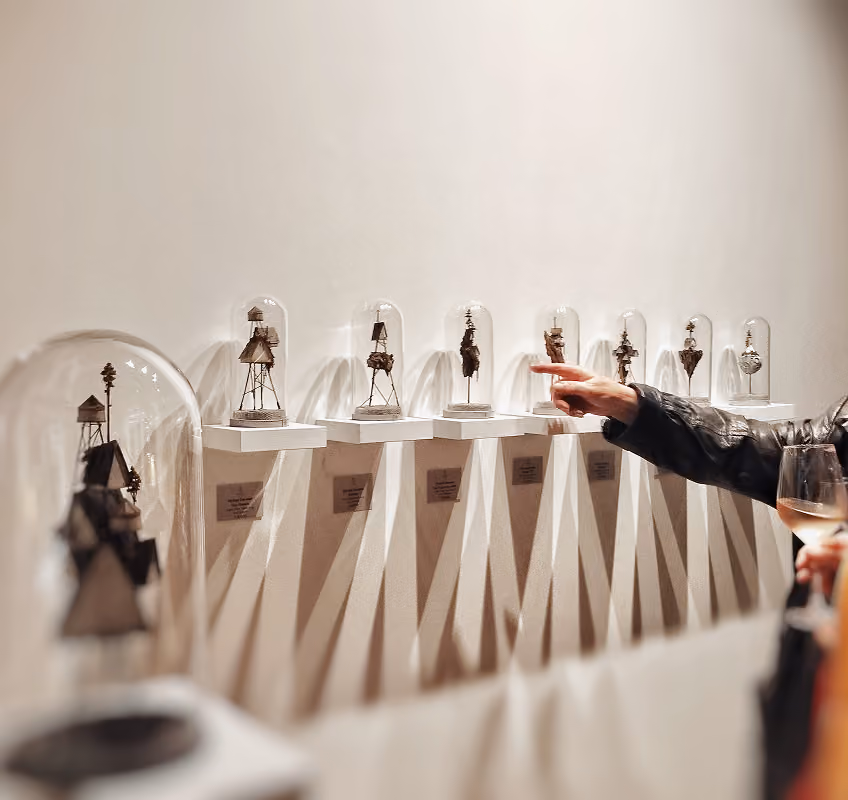 A person holding a glass of wine points at a series of small, intricate sculptures displayed under glass domes on a white wall, each mounted on individual white shelves with labels below them.