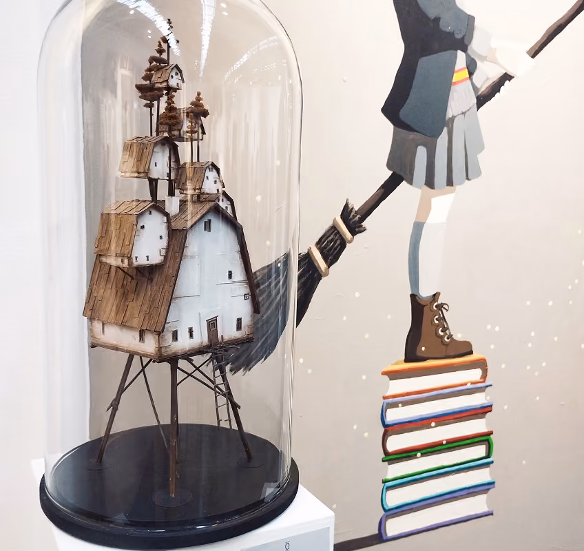 Miniature whimsical house model with multiple small houses on the roof, elevated on tall thin legs, displayed under a glass dome next to a wall painting of a person standing on a stack of colorful books holding a broomstick.