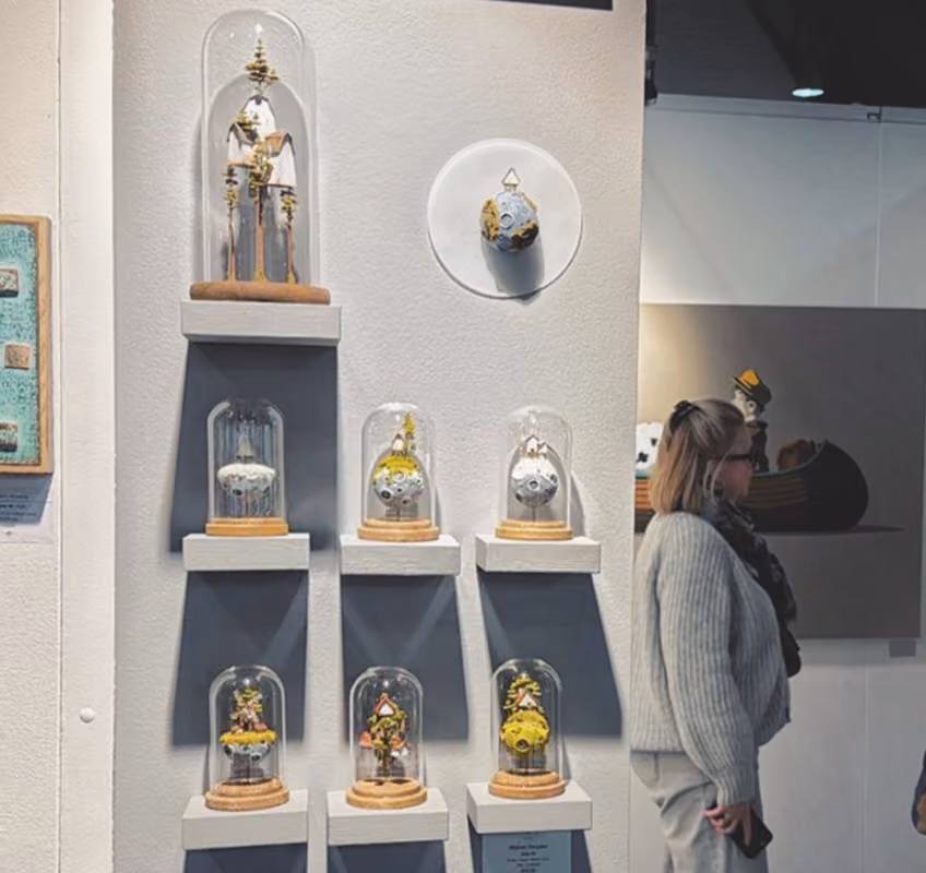 Wall display of nine miniature architectural models enclosed in glass domes, arranged in a grid on white shelves with shadows, a woman standing nearby viewing art in a gallery setting.