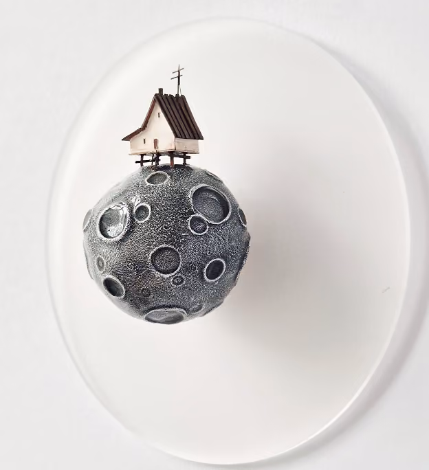 A small model of a textured cratered moon with a tiny house on stilts and an antenna on top, set against a plain white background.