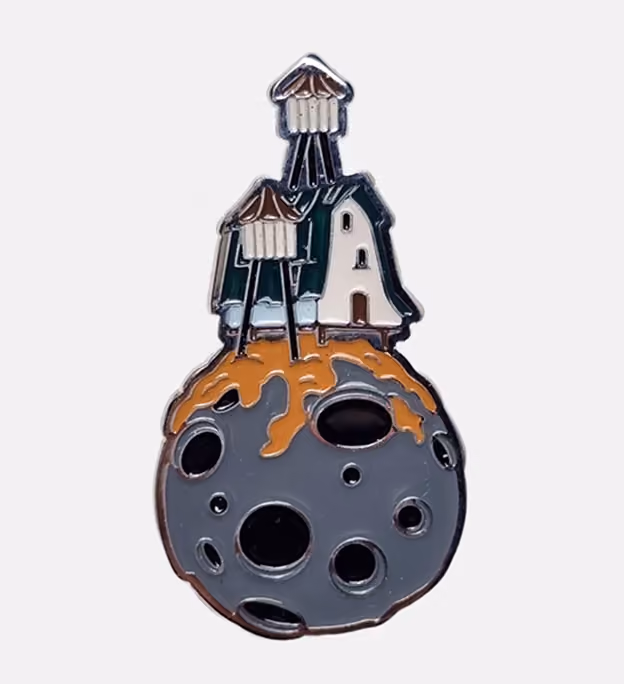 Enamel pin of a small white house with a dark roof and two stilts, positioned on top of a gray cratered moon with orange lava flowing around the base of the house.