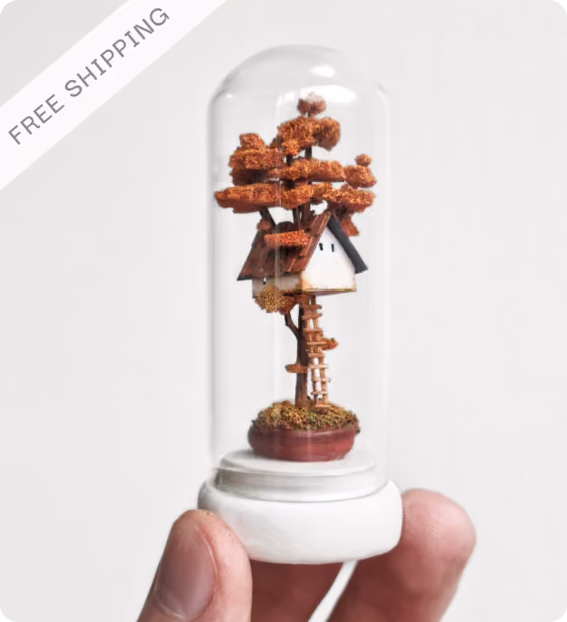 Hand holding a miniature treehouse with orange foliage and a small white house encased in a glass dome with a white base and a corner label reading 'FREE SHIPPING'.