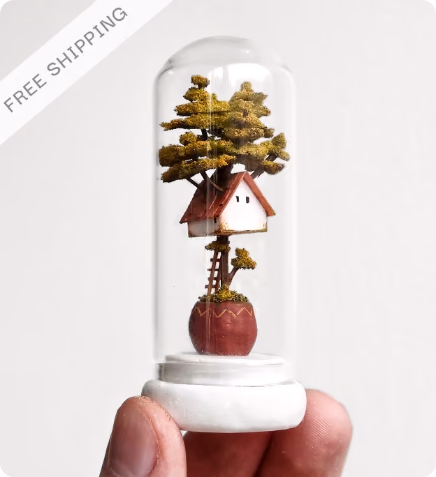 Hand holding a small glass dome encasing a miniature treehouse with green foliage, a ladder, and a round red base, on a white background with a free shipping label in the top left corner.