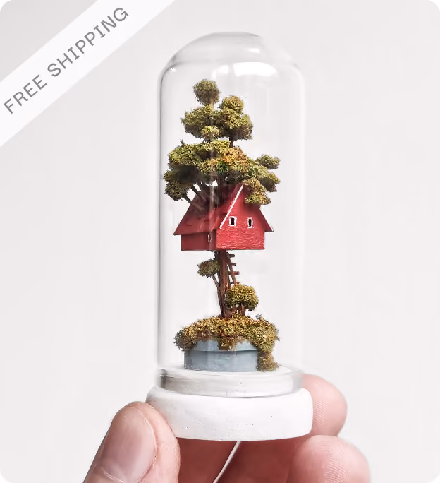 Hand holding a small glass dome with a miniature treehouse inside, featuring a red house elevated by a tree with green moss and a tiny ladder.