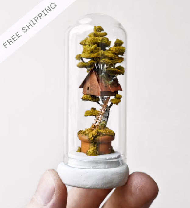 A person's fingers holding a small glass dome encasing a miniature tree with green foliage and a tiny wooden treehouse with a ladder in a small pot.