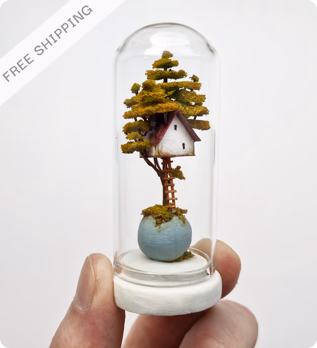 Hand holding a miniature treehouse inside a glass dome with a white base, featuring a tree with green foliage growing from a round blue pot and a small white house with a brown roof nestled among the branches.