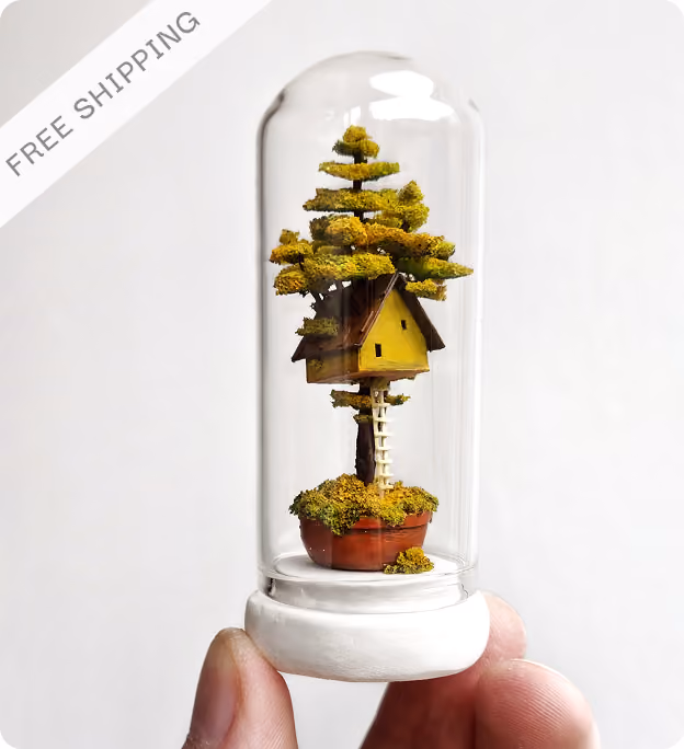 Hand holding a miniature treehouse inside a glass dome with a white base, featuring a tree with green foliage growing from a round blue pot and a small white house with a brown roof nestled among the branches.