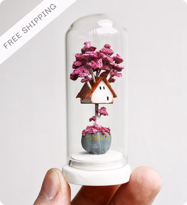 Hand holding a miniature treehouse inside a glass dome with a white base, featuring a tree with green foliage growing from a round blue pot and a small white house with a brown roof nestled among the branches.