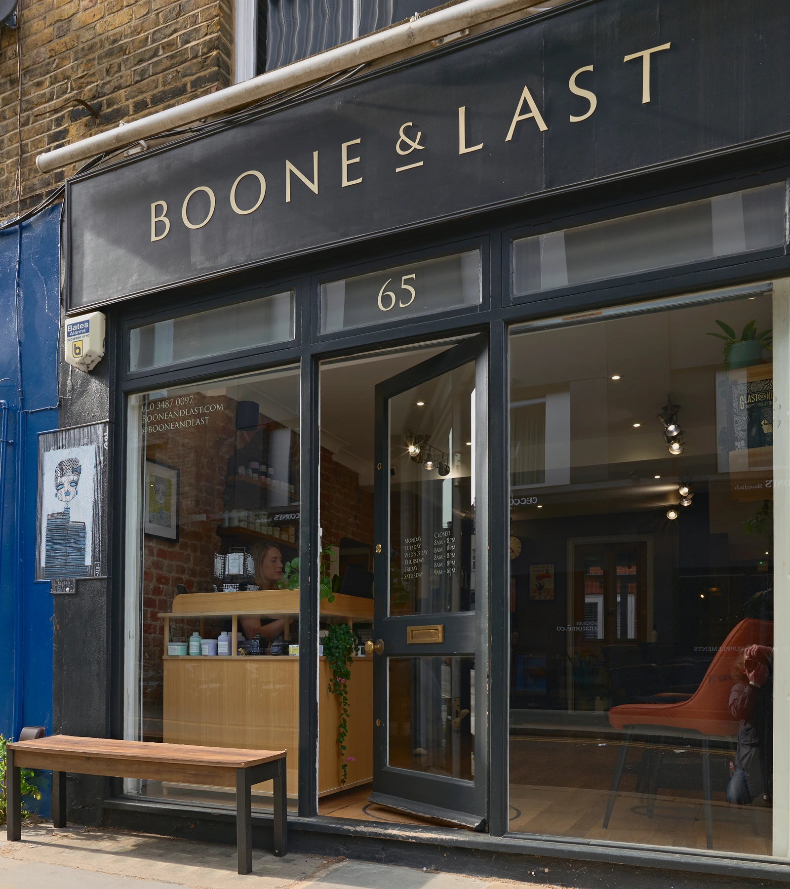 Front entrance of Boone & Last with open black door, wooden bench outside, and interior view of a brick-walled shop with shelves and a counter.