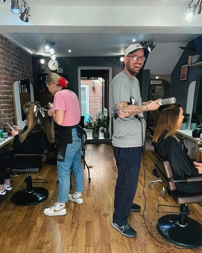 Two hairstylists working on clients in a sleek salon with wooden floors and exposed brick walls.
