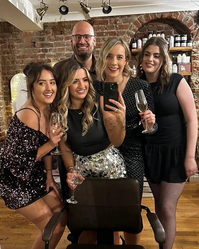 Group of four women and one man posing for a mirror selfie, with some holding glasses of wine, in a room with exposed brick walls and shelves of bottles.