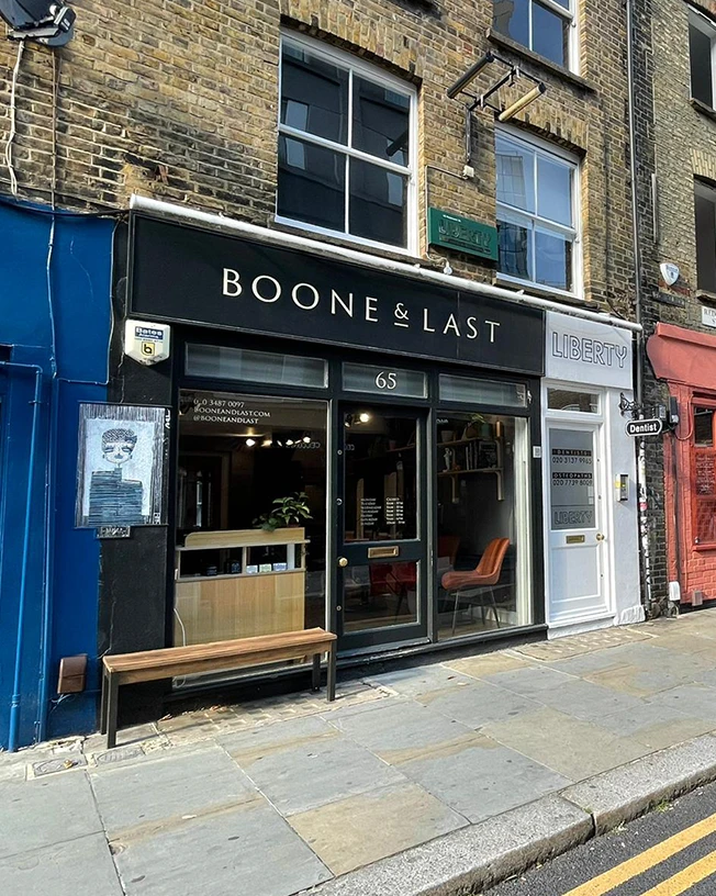 Street view of Boone & Last storefront with black facade and a wooden bench outside, next to Liberty and Dentist shops.