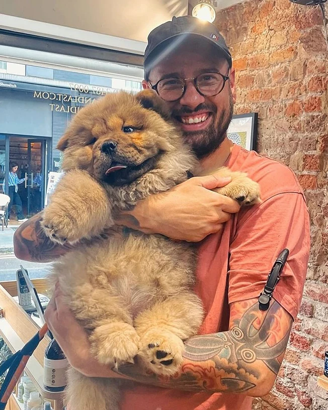 Man wearing glasses, a cap, and a coral t-shirt holds a fluffy chow chow puppy inside a cozy room with a brick wall.