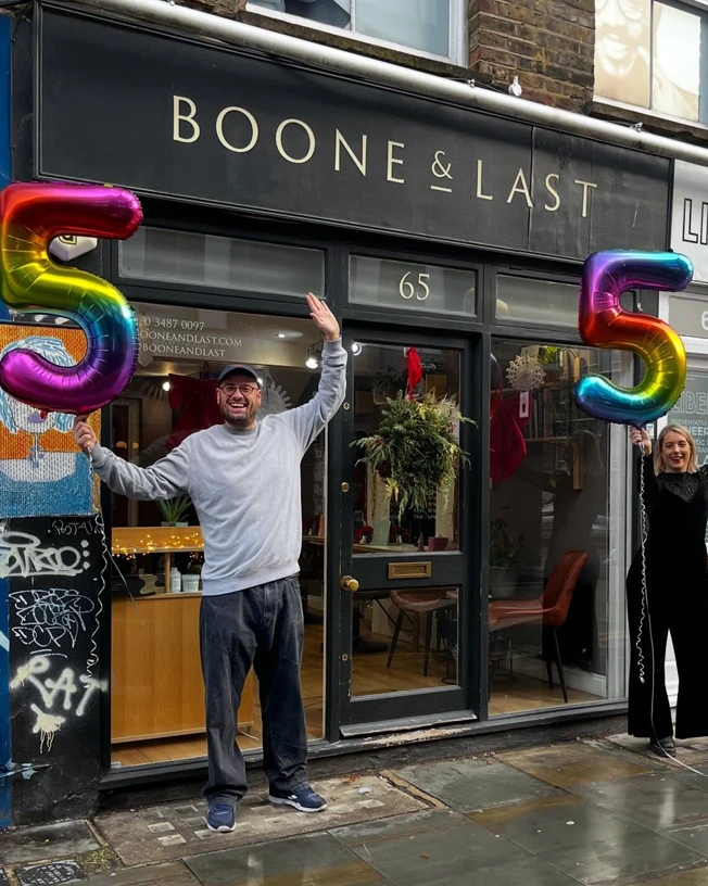 Two smiling people stand outside BOONE & LAST storefront holding colorful number 5 balloons.