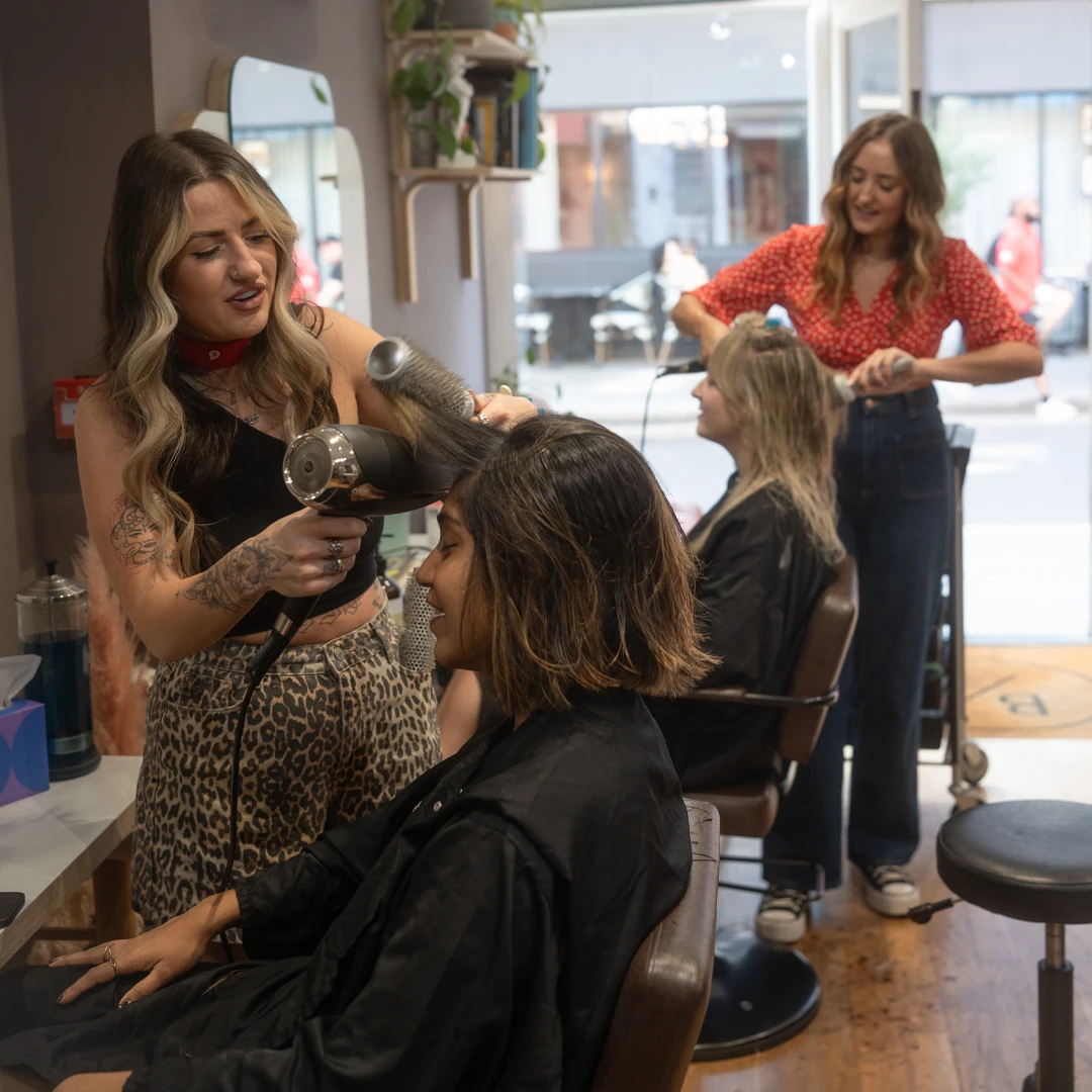 Two hairstylists blow-drying hair of two seated clients in a bright salon.