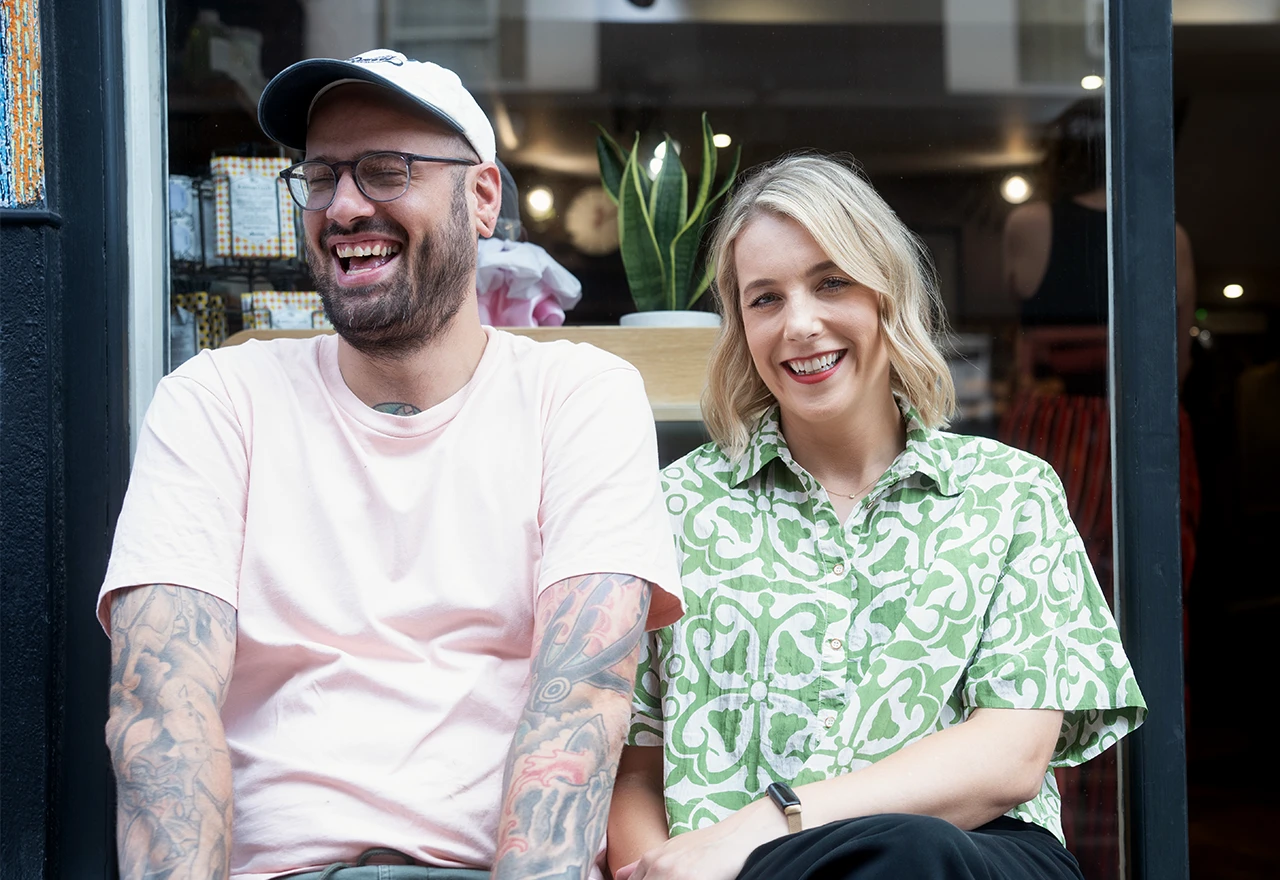 Smiling man with tattoos, glasses, and cap sitting beside a smiling woman in a patterned green shirt in front of a shop window with a potted plant inside.
