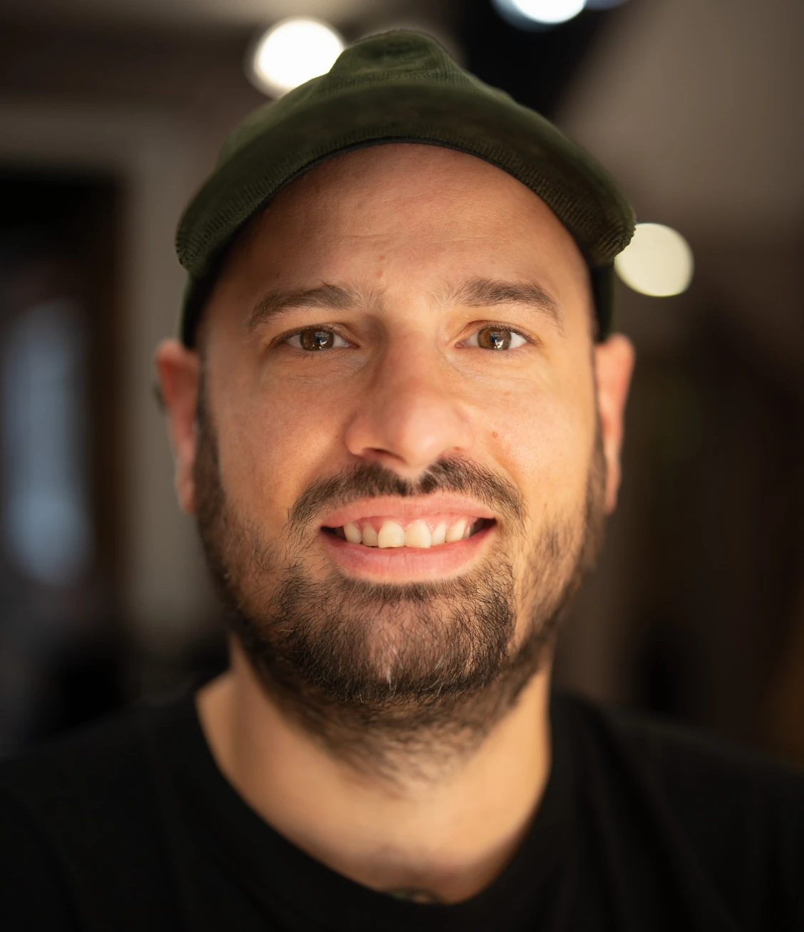 Smiling man with a trimmed beard wearing a dark green cap and black shirt in soft indoor lighting.
