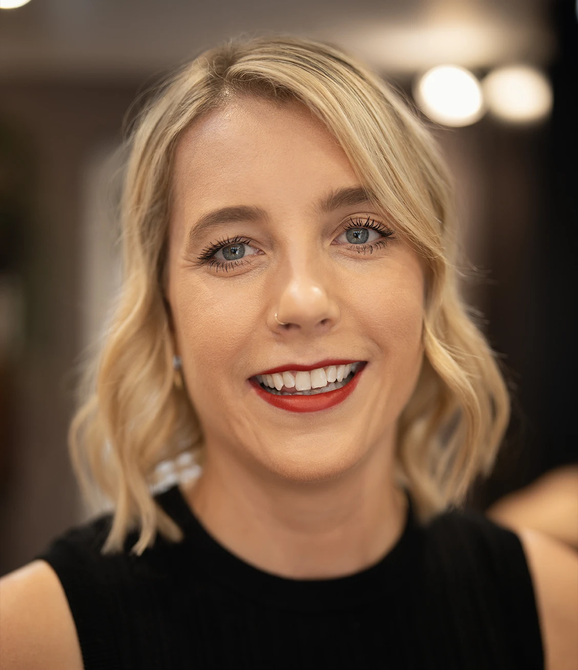 Close-up of a smiling woman with blonde wavy hair, blue eyes, and red lipstick wearing a black top.