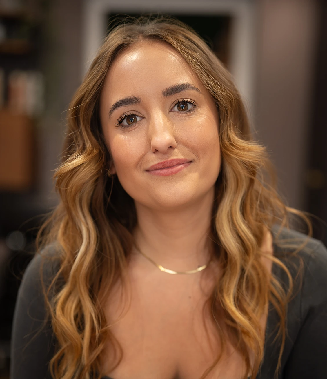 Portrait of a young woman with long wavy light brown hair wearing a dark top and a thin gold necklace, smiling softly.