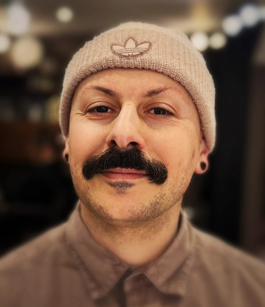Man wearing a beige beanie with an Adidas logo, a brown shirt, and a thick mustache with a small patch of hair below the lower lip.