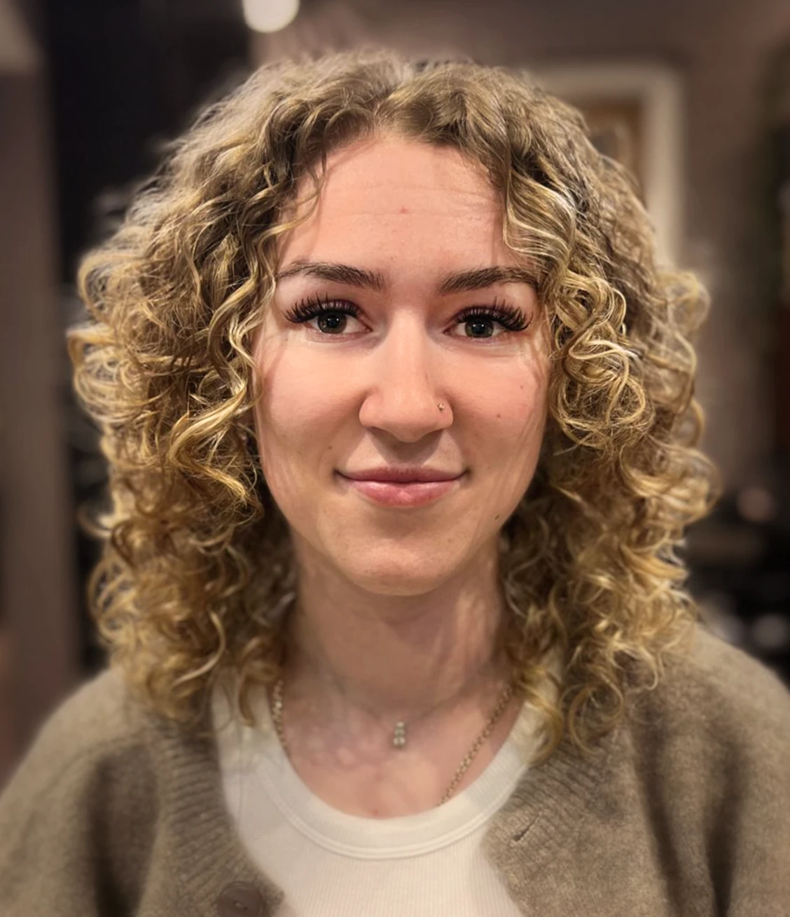Smiling woman with curly blonde hair wearing a black top and gold necklaces standing in front of green plants.