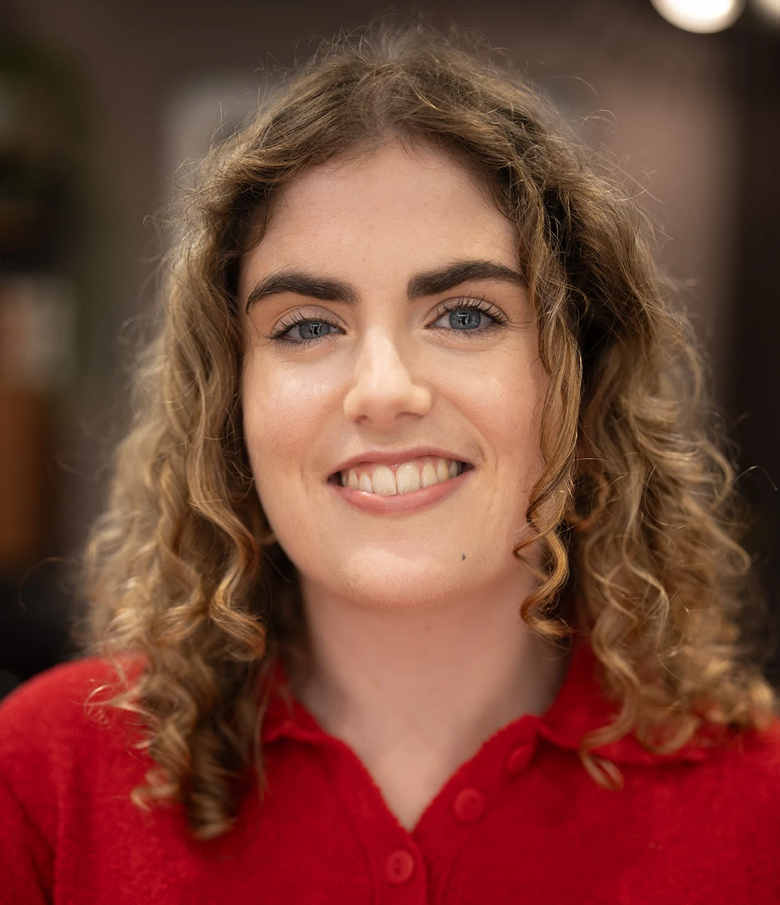 Smiling woman with curly blonde hair wearing a red buttoned shirt in a softly blurred indoor setting.