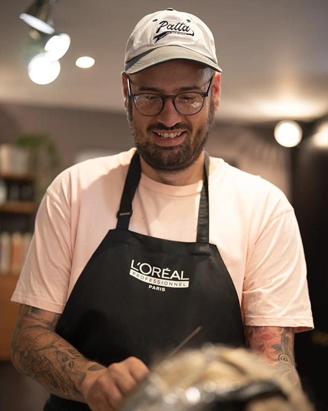 Smiling man with glasses and tattoos wearing a Patta cap and a L'Oréal Professionnel Paris apron.
