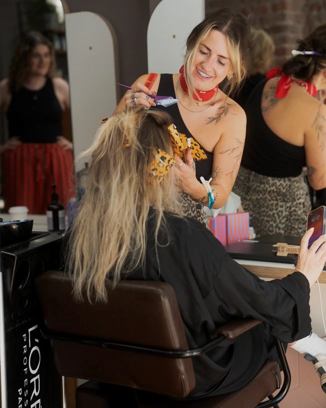 A hairstylist with tattoos and a red bandana dyeing a seated client's hair in a salon.